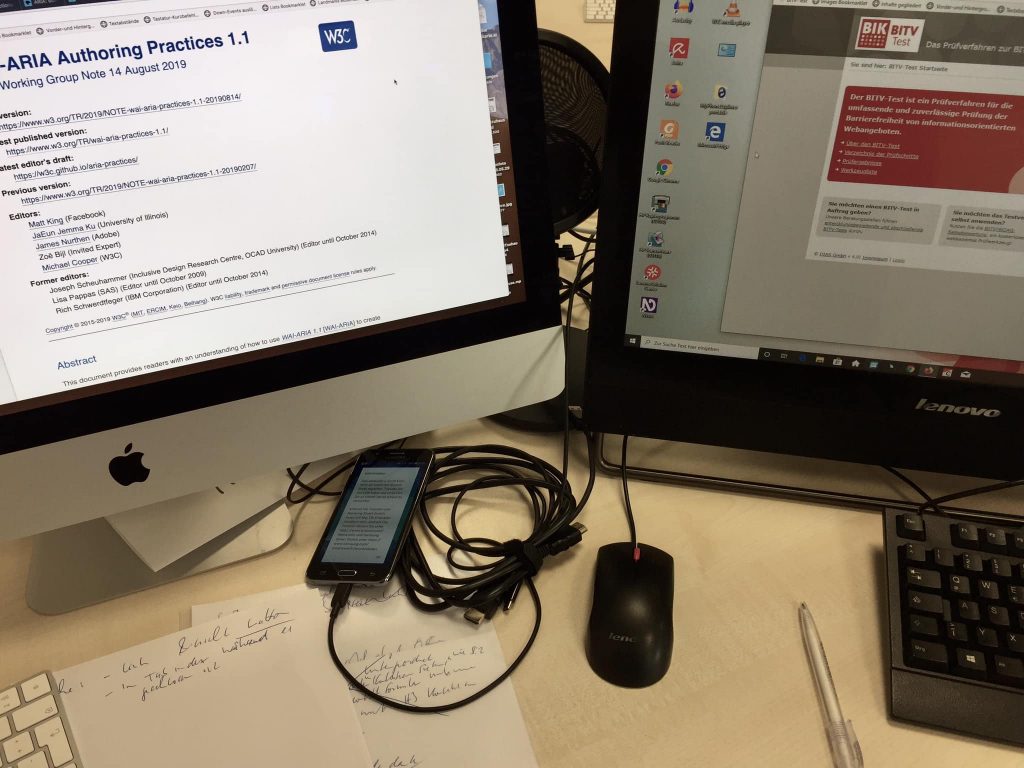 Arbeitsplatz bei PIKSL zum Testen von Internetseiten. Das Bild zeigt zwei Monitore mit Internetseiten, ein Smartphone und handbeschriebene Zettel.